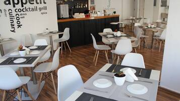 Un interior de restaurante con mesas dispuestas para comer, una zona de bar y una nube de palabras en la pared.