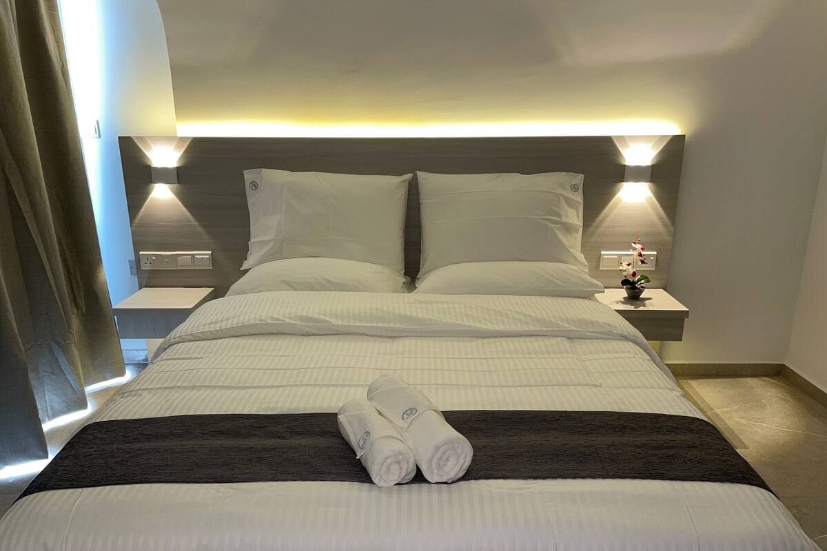 A neatly made bed with white linens, two pillows, and a folded towel on the bed. There is a nightstand with a lamp and a small potted plant beside the bed.