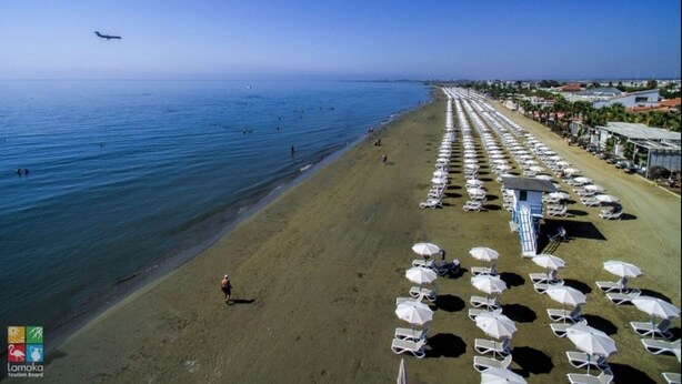 Une plage bordée de rangées de chaises longues et de parasols, un ciel dégagé et un avion qui vole au loin.
