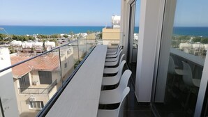 Un balcon avec une longue table et des chaises blanches, donnant sur une zone côtière avec des bâtiments et un ciel dégagé.
