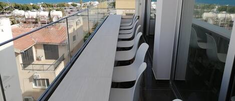 Un balcon avec une longue table et des chaises blanches, donnant sur une zone côtière avec des bâtiments et un ciel dégagé.
