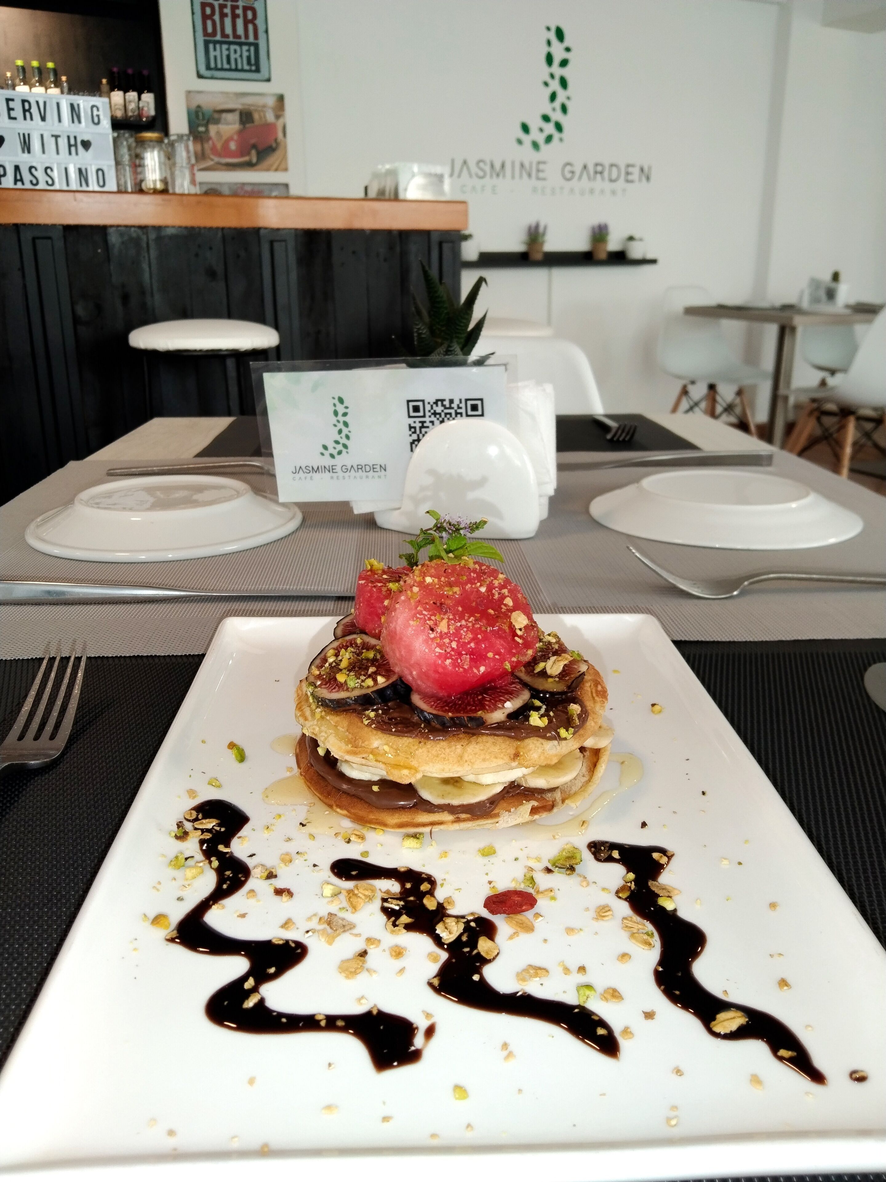 Ein Dessert auf einem weißen Teller mit Schokoladensauce und Nüssen, ein Schild für Jasmine Garden und ein Barbereich im Hintergrund.