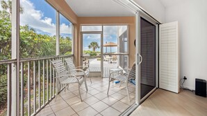 Un balcon avec des chaises blanches et une table, donnant sur un patio agrémenté d’un palmier et d’un parasol de plage.