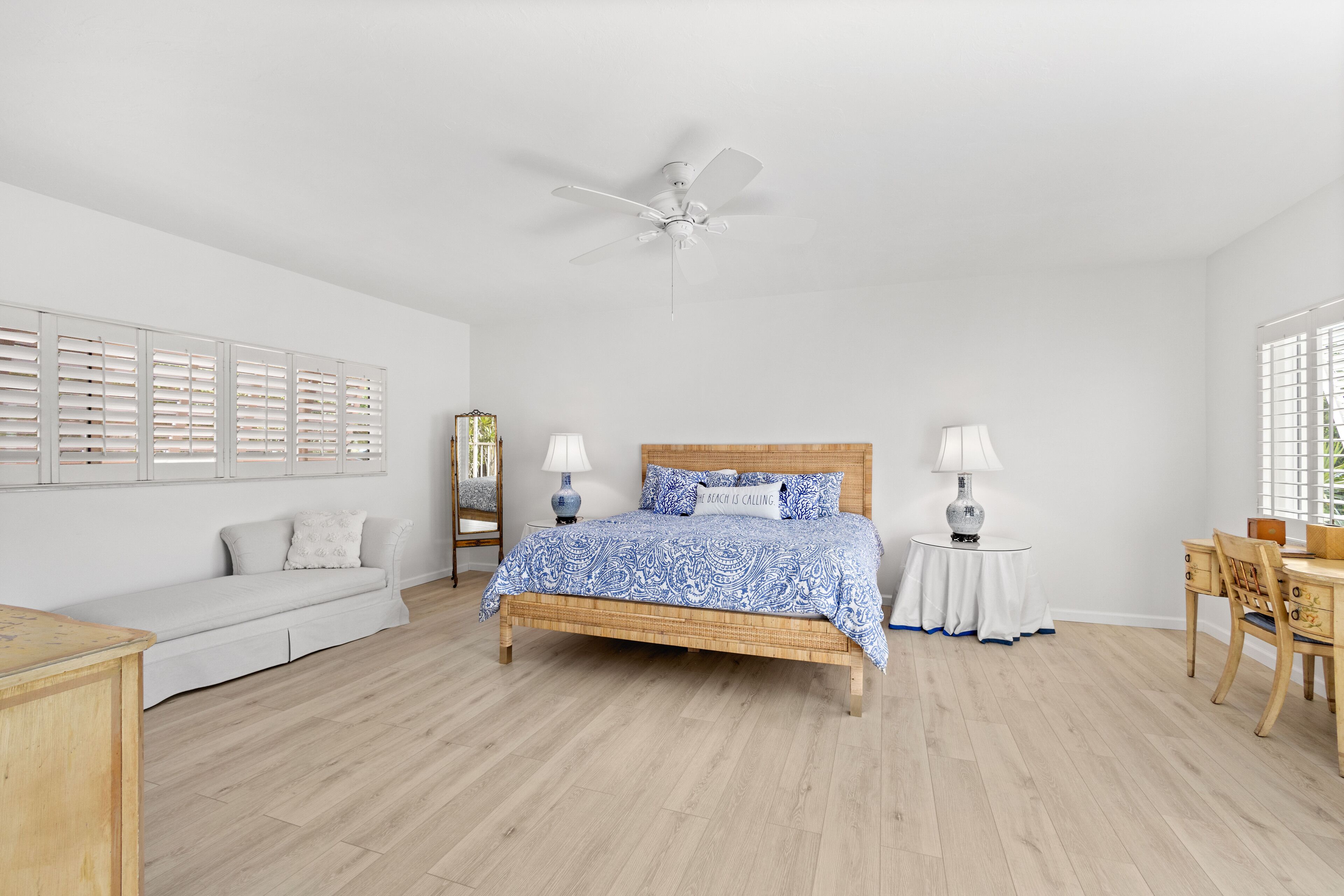 Une chambre à coucher comprenant un lit, un ventilateur de plafond, une table de chevet, une lampe, une chaise, un canapé et une fenêtre avec des stores.