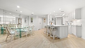 Une cuisine moderne avec des meubles blancs, un îlot central doté de tabourets de bar et un coin repas avec des tables à plateaux en verre et des chaises blanches.