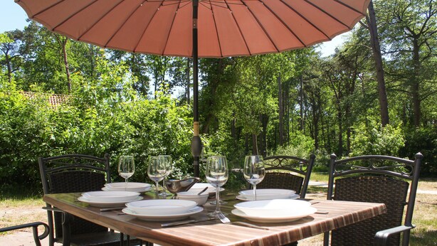 Une table à manger installée à l’extérieur, avec des assiettes, des verres à vin et des couverts, entourée de chaises en osier sous un grand parasol.