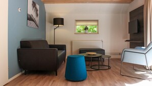 Un salon avec un canapé, un pouf bleu, une petite table sur laquelle se trouve un bol, une lampe sur pied, une fenêtre avec une plante et un téléviseur fixé au mur.