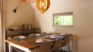 Un coin repas avec une table en bois dressée pour quatre personnes, un luminaire suspendu, une fenêtre donnant sur la verdure et un petit meuble contenant un téléphone et un cadre photo.