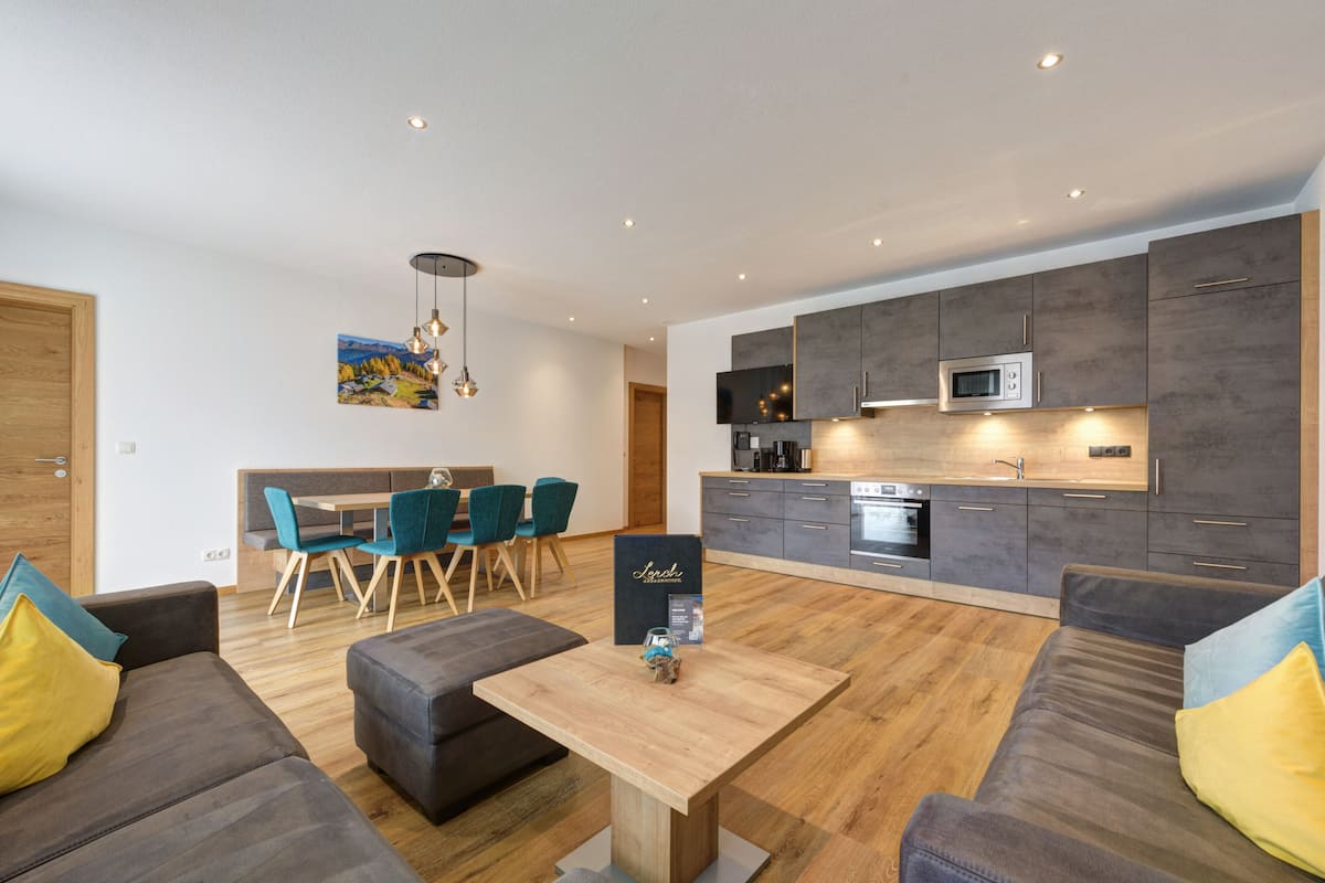 Ein modernes Wohnzimmer mit Essbereich, grauen Sofas, einem Holztisch und einer Küche mit grauen Schränken und einer Mikrowelle.