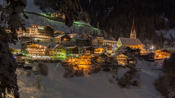 Un pueblo cubierto de nieve con edificios iluminados, incluyendo una iglesia con una alta aguja, y una pista de esquí al fondo.