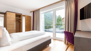 Habitación de hotel moderna con una cama grande, un armario de madera, un área de descanso con una silla morada y un balcón con vistas panorámicas a la montaña.