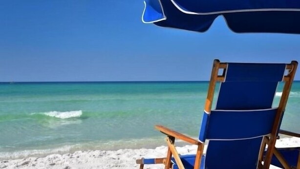 Una spiaggia con cielo azzurro, mare turchese e una sedia blu sotto un ombrellone blu.