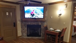 A living room with a stone fireplace, a television mounted above it, a dining table with chairs, and a leather couch.