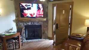 A living room with a stone fireplace, a television mounted above it, a dining table with chairs, and a leather sofa.