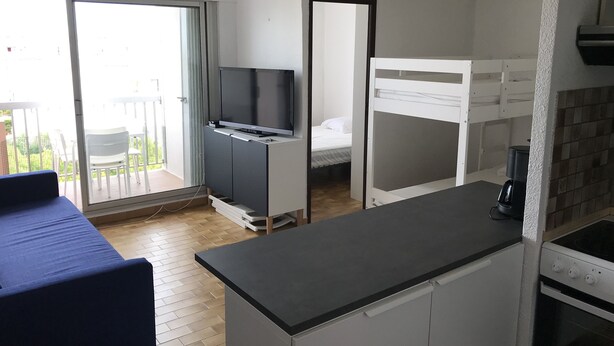 Une chambre compacte comprenant un coin cuisine, un lit, une télévision et un balcon aménagé avec du mobilier d’extérieur.