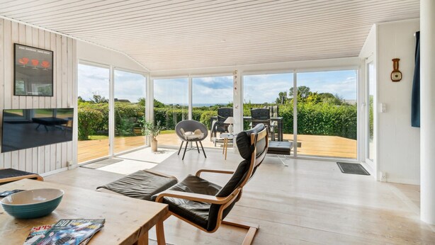 Un espacio de sala de estar moderno y abierto con un amplio ventanal corredera que da a una terraza de madera. En el interior, hay zonas de estar cómodas con sillones de cuero y una mesa de comedor.