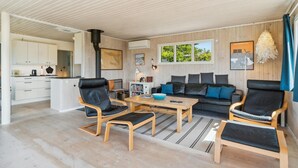 Una sala de estar moderna con un sofá, dos sillones, una mesa de centro y una zona de cocina con armarios blancos y una estufa.