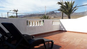 Una terraza en la azotea con suelo de baldosas, una pared blanca con marcos de ventanas decorativos y vistas a montañas y aerogeneradores a lo lejos.
