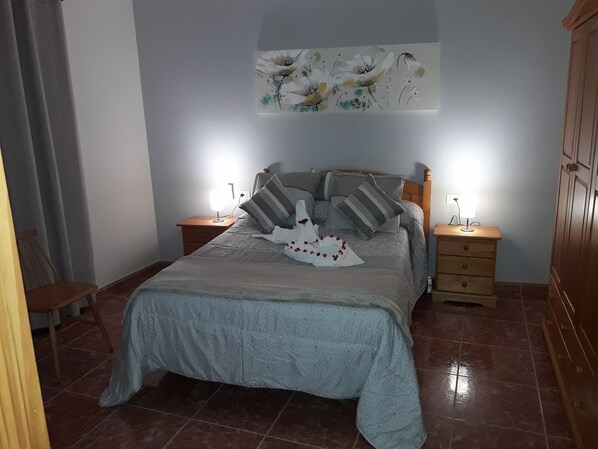 A bedroom with a bed, two bedside tables, a nightstand, a wooden wardrobe, and a wall-mounted artwork.