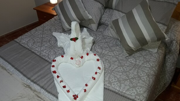 Una cama hecha con esmero, una toalla con forma de corazón y flores rojas, ropa de cama con estampado gris y almohadas a rayas.