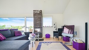 Een moderne woonkamer met een grijze bank, een glazen salontafel, een open haard, een televisie en een groot raam met uitzicht op groen.