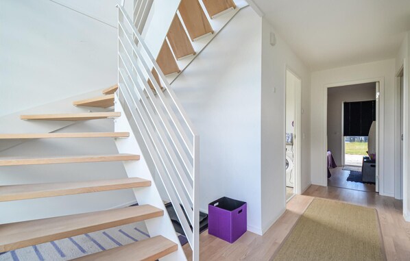 Un escalier moderne avec des marches en bois et des rampes blanches, menant à un couloir où se trouve une boîte de rangement violette et d’où l’on peut admirer un espace extérieur.