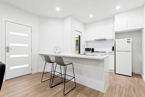A modern kitchen with white cabinetry, a central island with bar stools, a clock, and a refrigerator. - Domi Rentals - The Hampton Homes (Moorabbin)