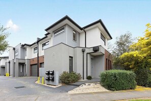 A modern building with a flat roof, large windows, and a parking area with yellow bollards. - Domi Rentals - The Hampton Homes (Moorabbin)