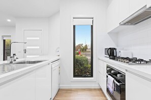 A modern kitchen with white cabinetry, a built-in oven, a gas stove, and a window with a view of the outside. - Domi Rentals - The Hampton Homes (Moorabbin)