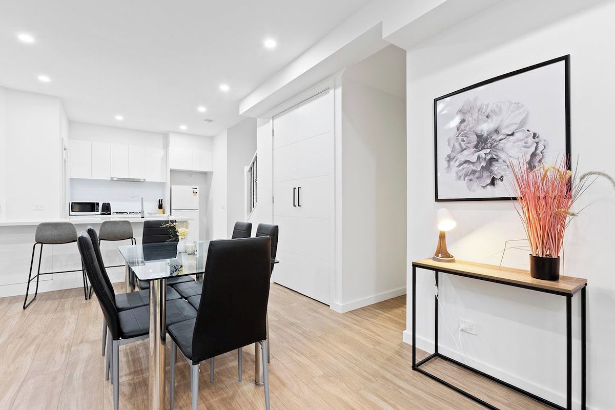 A modern kitchen with white cabinets, a dining area with a glass table and black chairs, and a framed artwork on the wall.