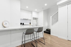 A modern kitchen with white cabinetry, a stainless steel range hood, and a built-in oven. There are two bar stools and a wall clock. - Domi Rentals - The Hampton Homes (Moorabbin)