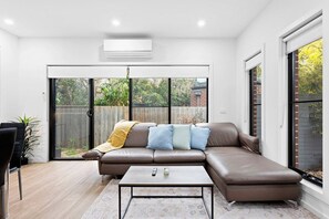 A modern living room with a leather sofa, a coffee table, and a sliding glass door leading to a backyard. - Domi Rentals - The Hampton Homes (Moorabbin)