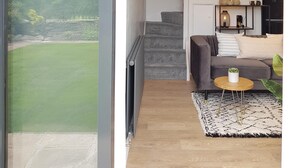 En moderne stue med en grå sofa, et rundt sofabord og udsigt til et grønt landskab gennem et stort vindue.