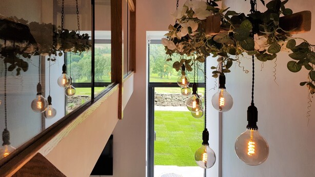 En trappe med hængende lyspærer og grønne planter, der fører op til en balkon med udsigt over en grøn græsplæne.