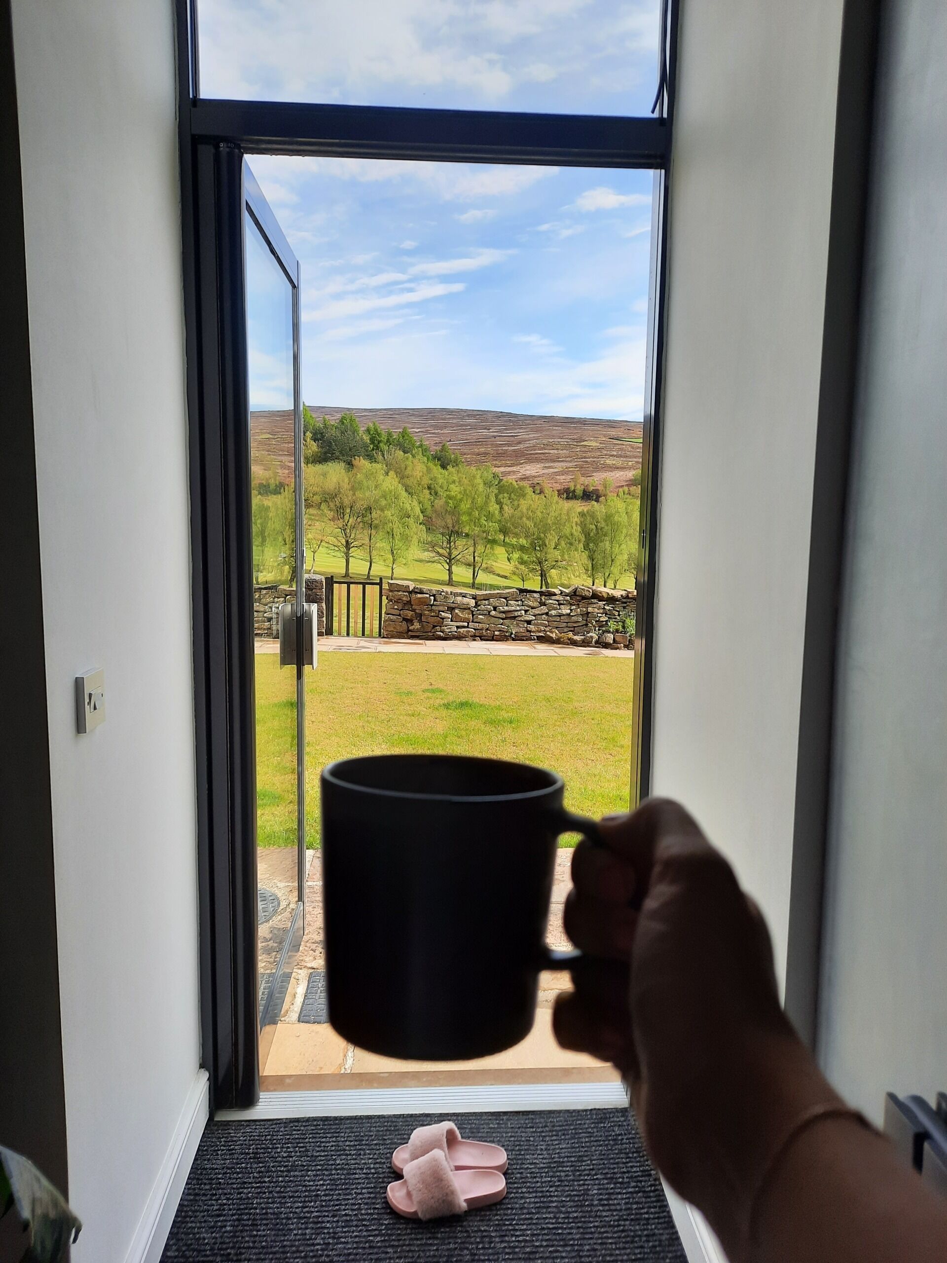 En hand håller i en svart mugg framför en dörr som leder ut till en utsikt över en gräsklädd äng och en stenmur.