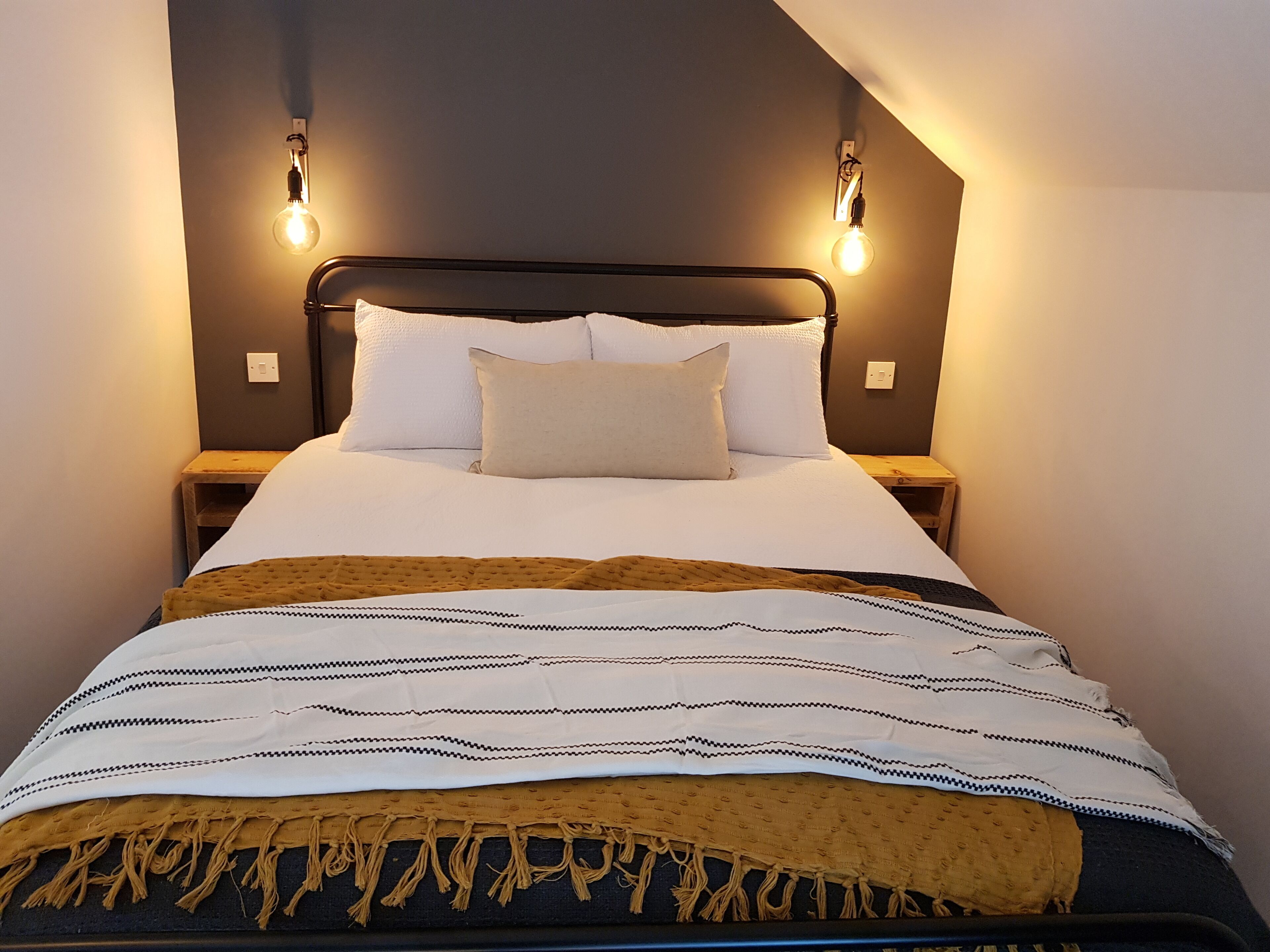 Una cama bien hecha con edredón blanco, dos almohadas y una manta decorativa estampada. Hay dos mesitas de noche con lámparas a cada lado de la cama.