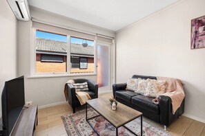 A living room with a black leather sofa, a wooden coffee table, a black armchair, a television, and a large window with a view of a brick building.