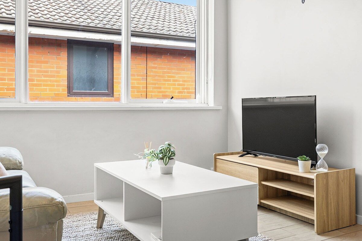 A modern living room with a white coffee table, a TV on a wooden stand, and a window overlooking a brick building.