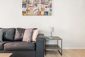 A modern living room with a black leather sofa, a small wooden side table, and a wall-mounted collage of travel photos.