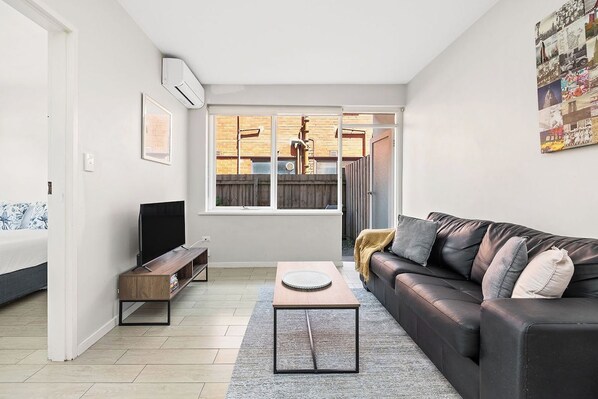 A modern living room with a sofa, a coffee table, a TV on a stand, and a window with a view of a brick wall and a fence.