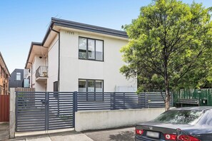 Un edificio residencial moderno de dos pisos con balcón, entrada con reja y un árbol en frente.