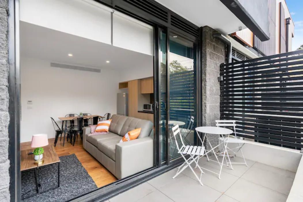 A modern living space with a sofa, dining table, and chairs, a small table with a lamp, and a balcony with a table and chairs.