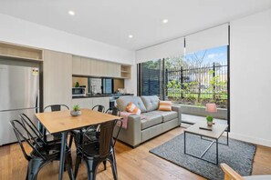 A modern kitchen with built-in appliances, a dining area with a wooden table and chairs, a grey sofa with cushions, and a view of a fenced garden through large windows.