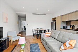 A modern living room with a grey sofa, a TV, a dining table with chairs, and a kitchen area with cabinets and a microwave.