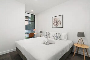 A modern hotel room with a large bed, a small table with a lamp, and a framed artwork on the wall.