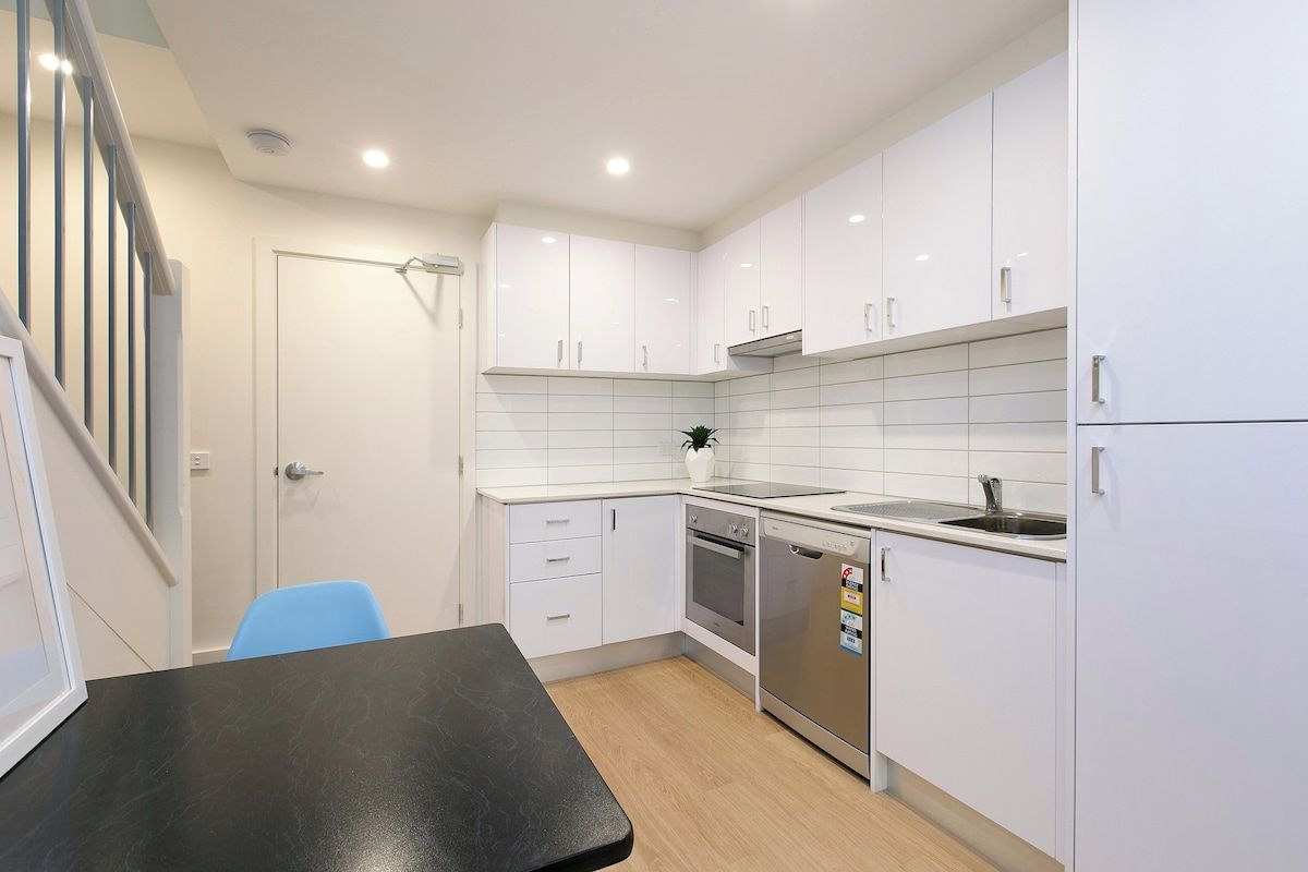 A compact kitchen with white cabinets, a black countertop, a dishwasher, and a blue chair.