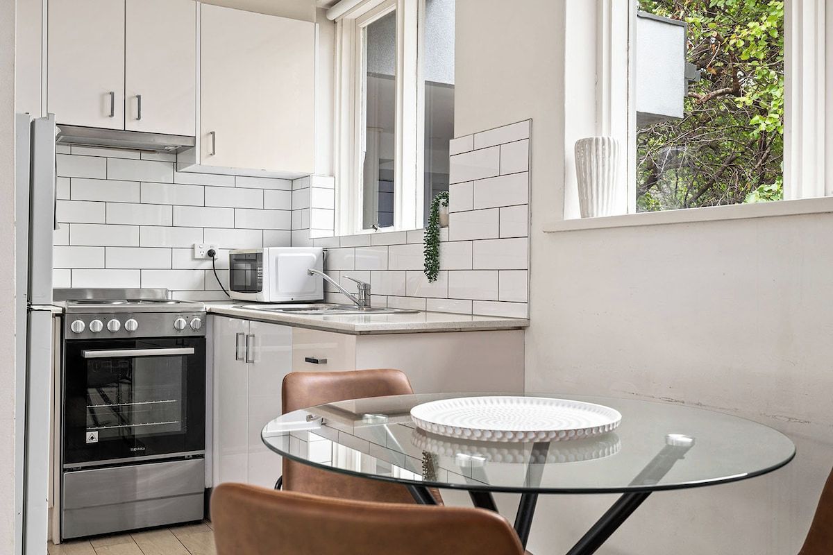 A modern kitchen with white cabinetry, a stainless steel oven, a microwave, and a glass table with chairs.