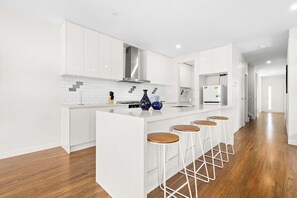 A modern kitchen with white cabinetry, a central island with stools, and a stainless steel range hood. - Domi Rentals - The Rayhur Residence (Clayton South)