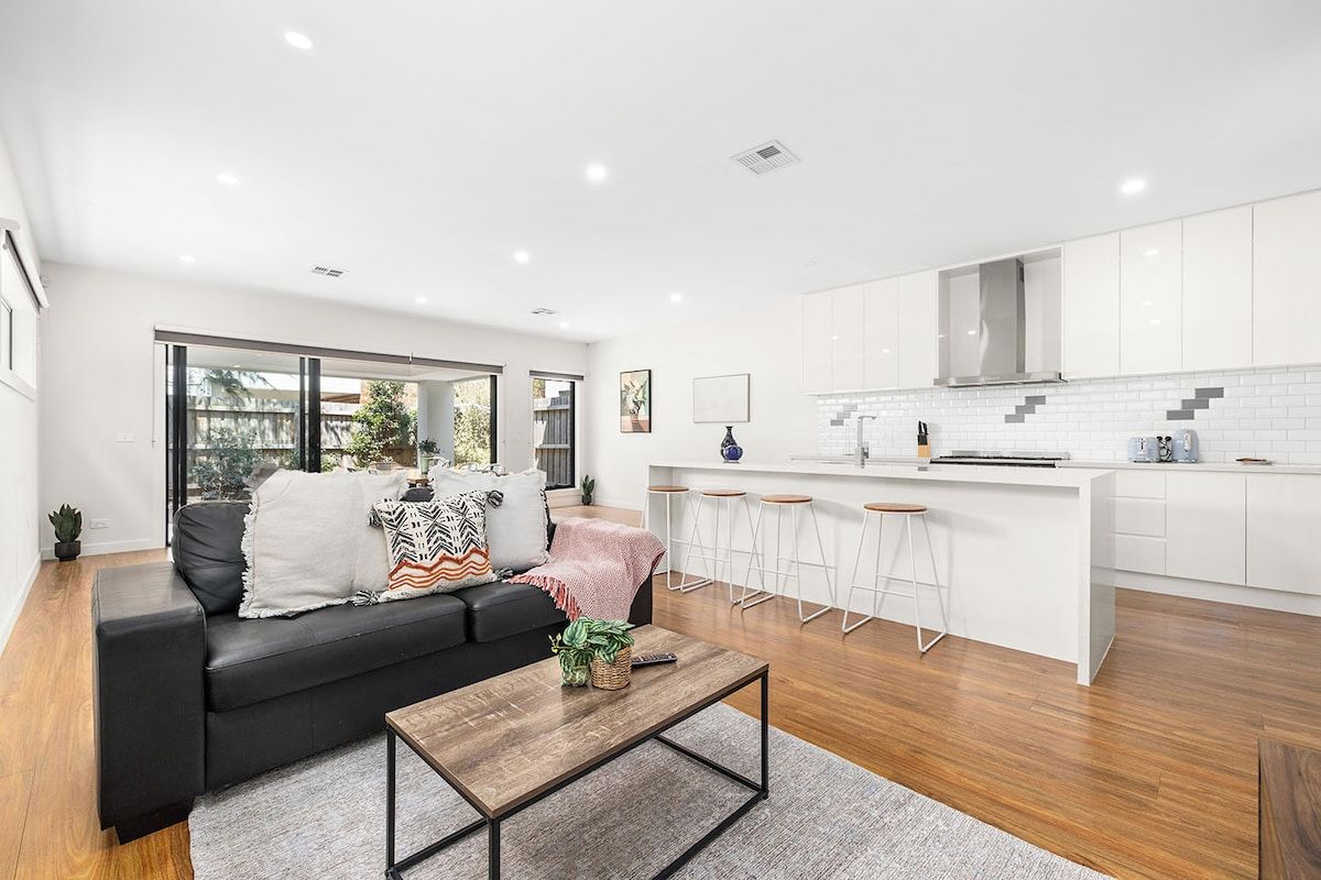 A modern living room with a black sofa, a wooden coffee table, and a kitchen area with white cabinets and a bar stool.