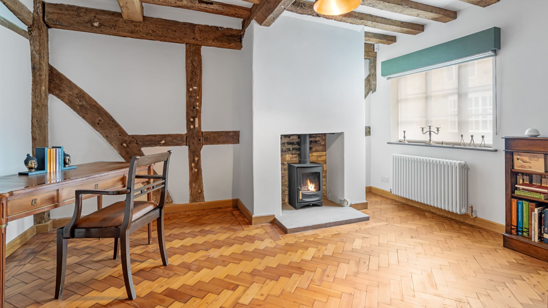 Une pièce avec des poutres apparentes, un parquet en point de Hongrie, une cheminée, un radiateur, un bureau avec une chaise et une bibliothèque.